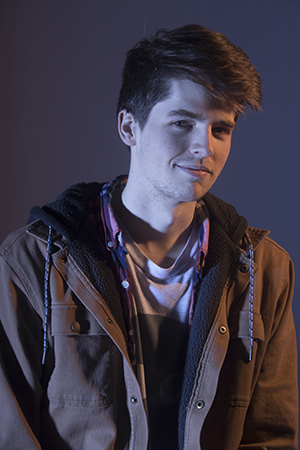 Austin Meros’s shoulders face forward looking straight on to the camera, lit dramatically, slight upturned smile upturned on the left side of the mouth