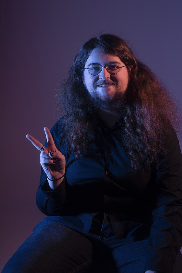 Brian flashes a peace sign at the camera and grins, he is wearing a black shirt and his nails are painted black. The lighting is a blue-ish purple and a salmon pink.