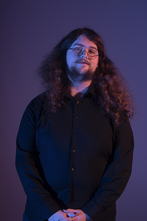 Brian clasps his hands in front of his body, looking confidently through his circular glasses. His hair is long and curly down to his biceps.