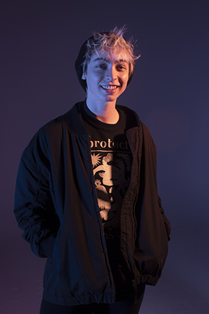 Cas stands facing the camera smiling, dramatic lighting, hands in jacket pockets, shirt obscured