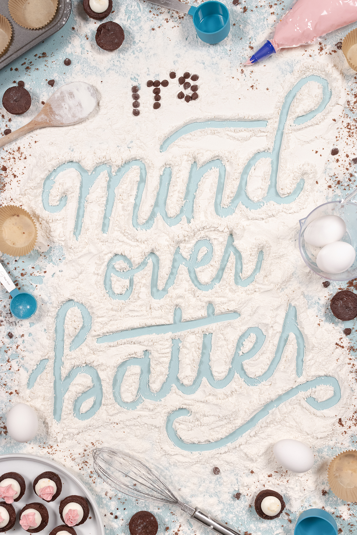 Hand-lettered script carved with a finger into flour, with ingredients and baking tools to make cupcakes designed into the image. Small chocolate cupcakes with pink and white frosting peek into the image. Script reads 'It’s mind over batter.'