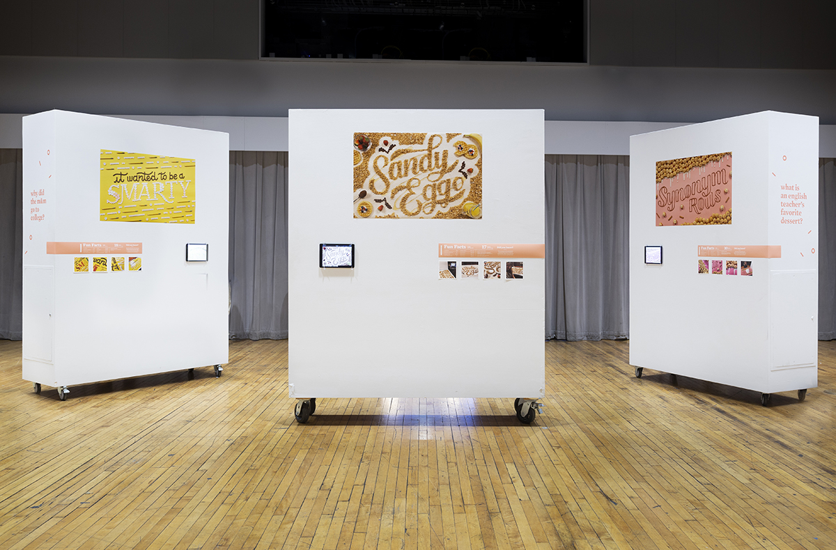 Photo from thesis gallery exhibit shows large posters of hand-lettered punch lines to jokes, made out of food. Small ipads and details of process are also installed with the large posters, vinyl hand lettering. Brightly lit, modern space and white moveable walls.