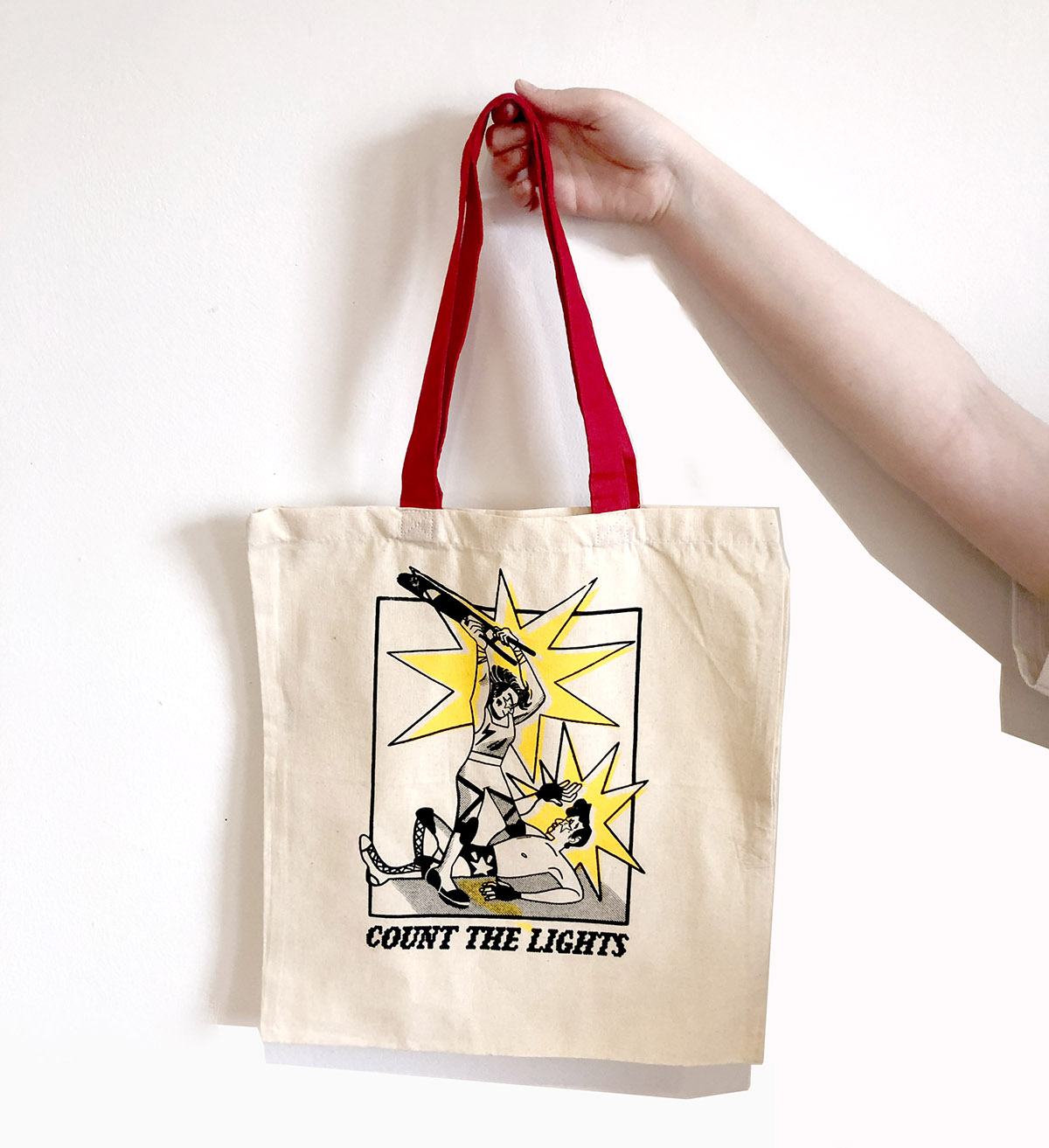 KD holds a tote bag with a red handle. Screen print on tote shows woman wrestler overtaking a man wrestler. Text below reads, 'count the lights'