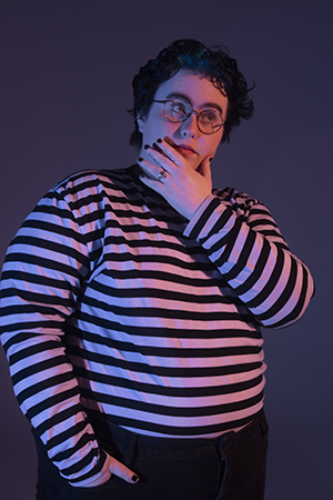 KD strands in dramatic lighting with right hand in pant pocket, left hand raised up to their chin, sideways gaze