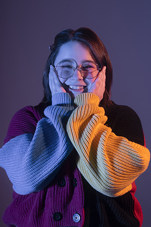 Lauren stands in dramatic lighting smiling at camera with hands cupping face