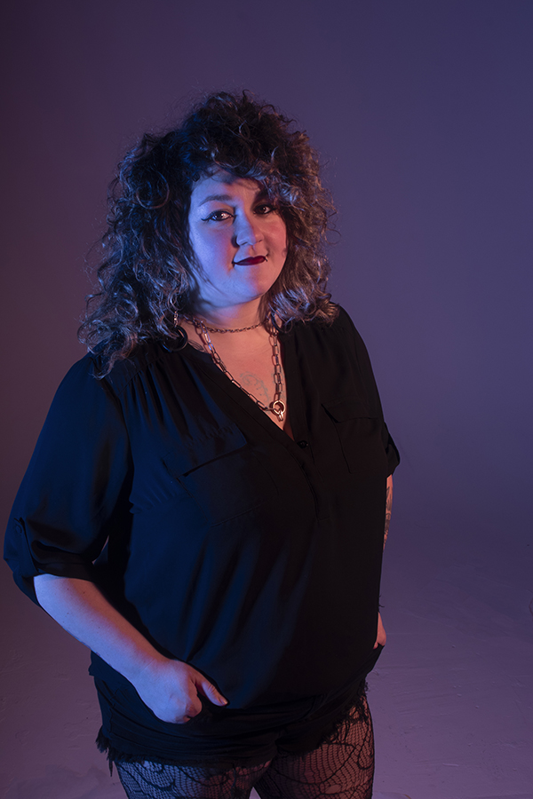 Mallary stands in dramatic lighting, with soft smile, turned to the right but looking at camera, with hands in her pockets.