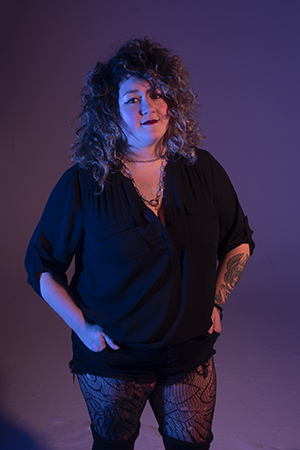 Mallary stands in dramatic lighting, with soft smile, facing front and camera directly, with hands in her pockets.