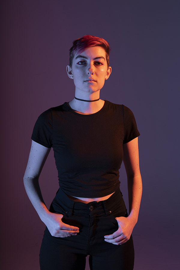 Tali stands confidently in dramatic lighting, hands in pockets, shoulders squared forward, eye contact forward