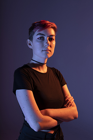 Tali stands confidently, with shoulders turned to her left, gaze to the camera, arms crossed at chest