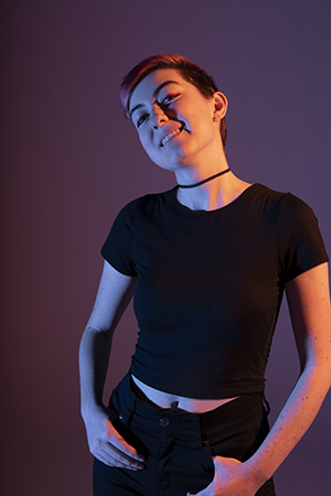 Tali stands confidently in dramatic lighting, hands in pockets, shoulders squared forward, eye contact forward, head tilted slightly back to her right, smiling