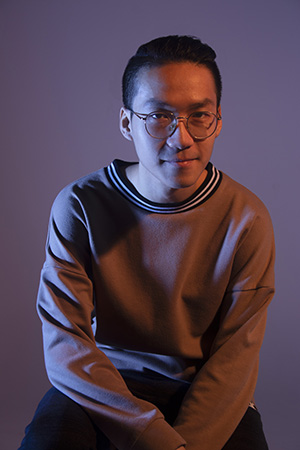 Trung sits in dramatic lighting, casually, one elbow resting on his knee, looking directly at the camera.