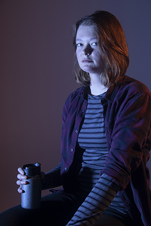 Zoe sits in dramatic cool lighting, serious gaze to camera, hands on lap with thermos in right hand resting on knee