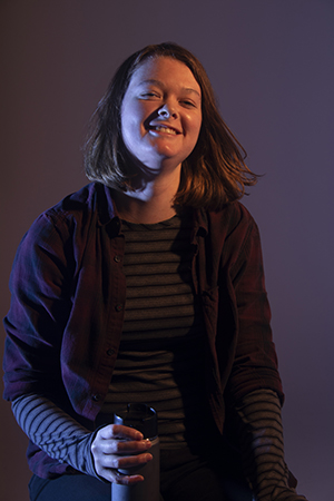 Zoe sits in dramatic lighting, thermos in right hand on knee, smiling with head tossing backwards
